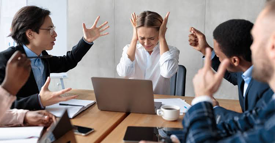 a group of people sitting around a conference table working with paper or laptops, with raised hands gesturing in frustration, and one woman with her hands over her ears